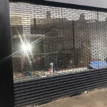 Black metal security shutter with circular punch holes and glazed panels.
